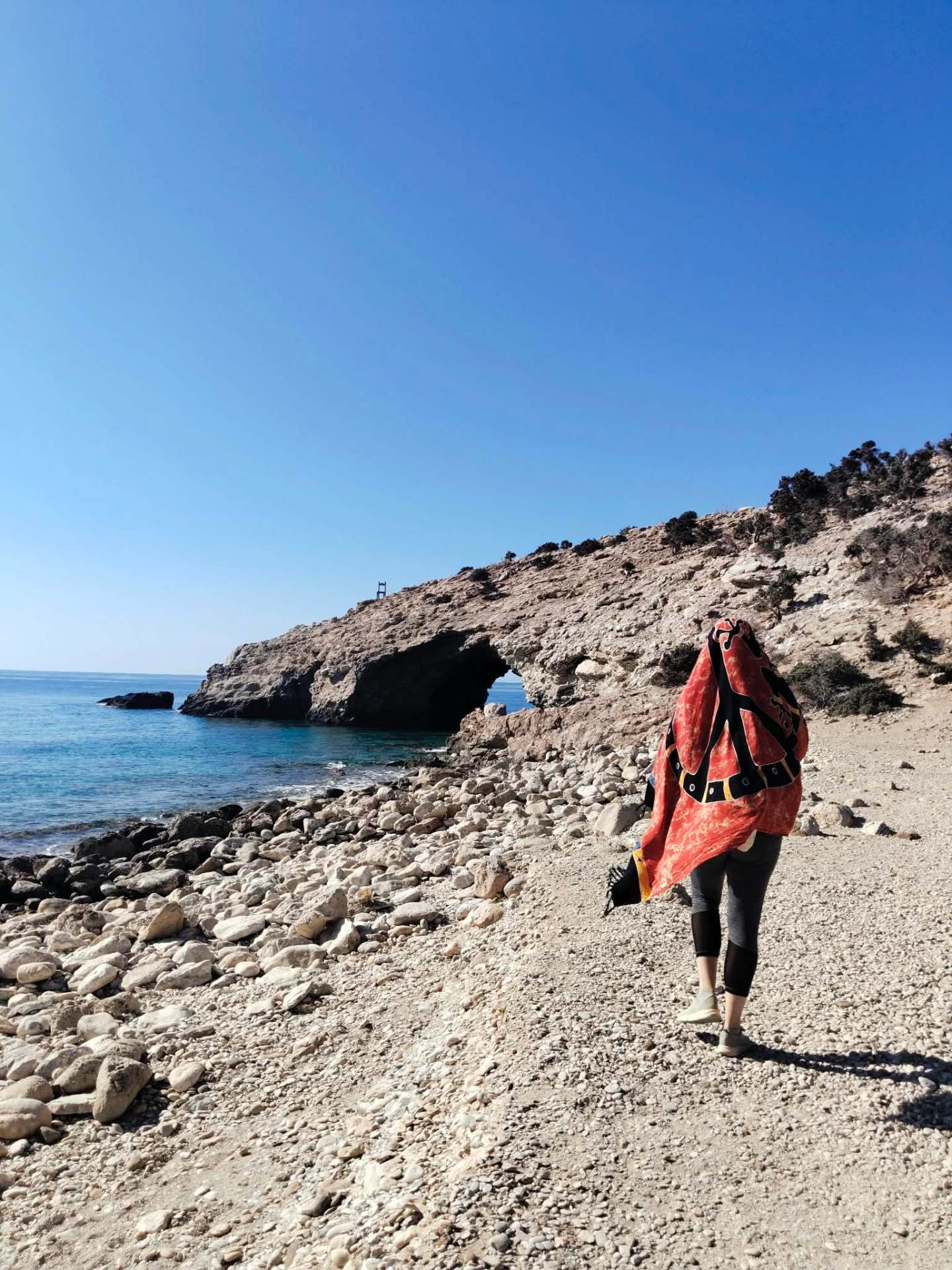 Lyu Gala caminando por una playa de piedras hacia el mar, envuelta en una tela roja con símbolo de paz